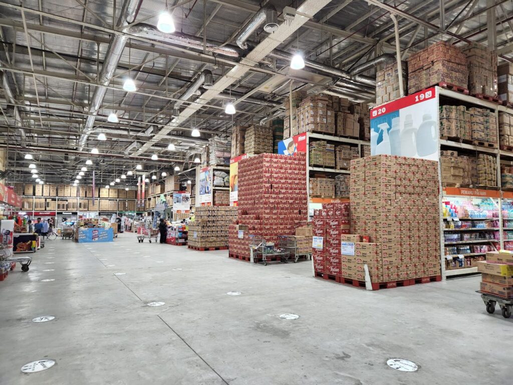 A large wholesale warehouse interior filled with stacked merchandise and shoppers browsing the aisles.